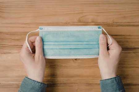 A person wearing a blue long sleeve shirt is holding a blue surgical mask with white straps on a...