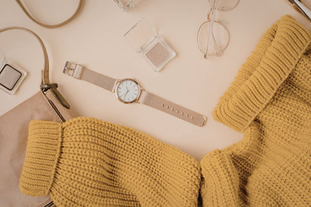 A stylish arrangement of autumnal accessories including a yellow sweater, a watch, glasses, and a purse on a beige background.の写真素材