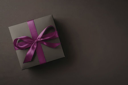 A gift box wrapped in dark paper with a purple ribbon tied in a bow on top, set against a dark...