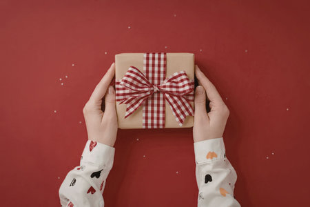 A pair of hands holding a gift box wrapped in brown paper and adorned with a red and white checkered...