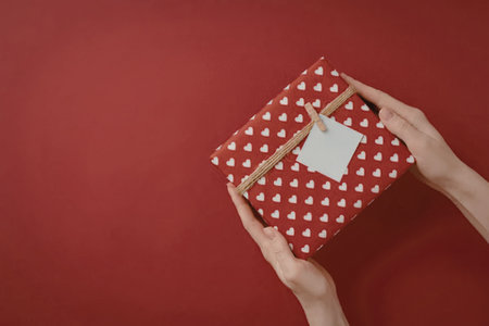 A pair of hands holding a red gift box with white polka dots and a brown ribbon on a red background.