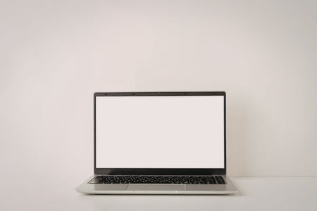 A laptop with a blank screen sits on a white surface against a white wall.
