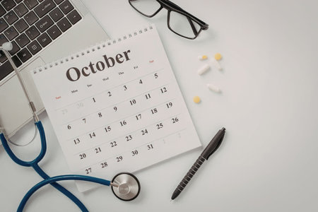 A calendar for October with a stethoscope, laptop, glasses, pen, and pills on a white surface.
