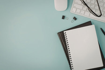 A light blue office desk with a spiral notebook, computer keyboard, mouse, glasses, paper clips, and...