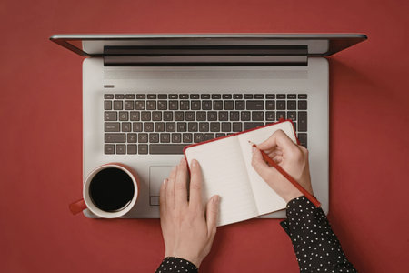 A person working on a laptop with a notebook and coffee on a red desk