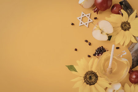 A Rosh Hashanah celebration setup featuring a honey jar with a dipper, sunflowers, apples, pomegranates, and a Star of David, all on an orange background.の写真素材