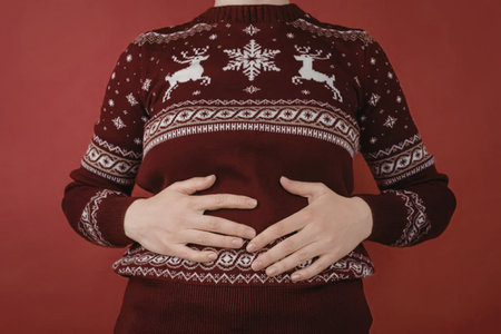 A woman wearing a red Christmas sweater with white reindeer and snowflakes, cradling her pregnant...