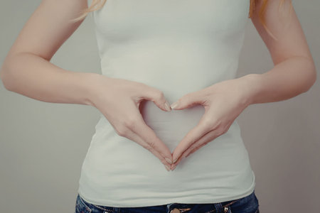 A woman wearing a white tank top and blue jeans making a heart shape with her hands around her...