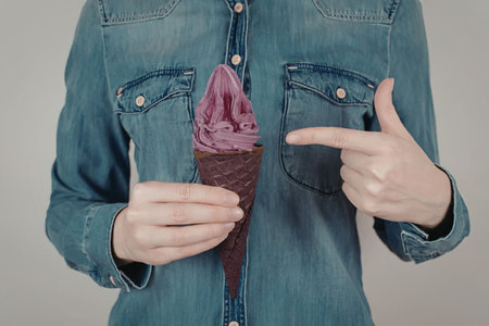 A woman in a denim shirt holding and pointing to a purple ice cream cone.