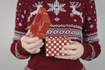 A person wearing a red Christmas sweater with white reindeer and snowflakes holds a red gift box with white polka dots and a red tissue paper.の写真素材