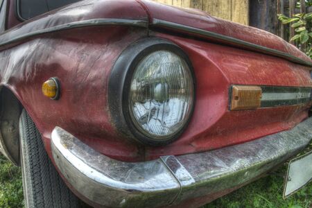 White headlight in a black frame of a red car with a silver bumper and a black wheel, reflections stand on the green grassの写真素材