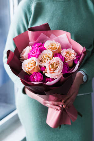 A girl standing by the window in a sea-green dress holds a burgundy bouquet with white and pink roses in her handsの写真素材