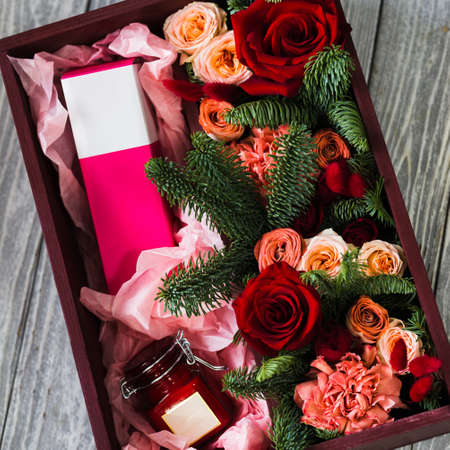 A burgundy wooden box with red roses, branches of a green Christmas tree, a pink perfume box in a glass jar lies on a wooden gray backgroundの写真素材