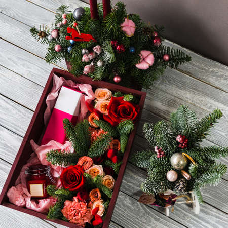 A burgundy wooden box lies on a gray wooden board inside which lies red roses with parts of the foliage of a Christmas tree with a pink perfume box and a glass jarの写真素材