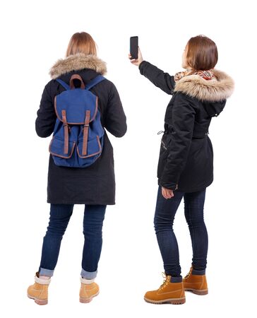 Back view of two young woman photographed on a mobile phone in winter jacket. Rear view people collection. backside view of person. Rear view. Isolated over white background.の写真素材