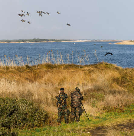 two men in camouflage observe aquatic birds near a swampの写真素材