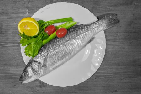 Color isolation effect of white dish with sea bass, lemon, celery and tomatoes on wooden background. Concept: cuisine based on fishの写真素材