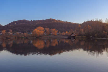 Hills reflected in the waters of a lake with a hut on its banks at sunsetの写真素材
