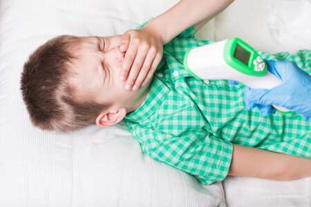 The boy in bed coughs, covering his mouth with his hand, and he measures the temperature