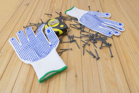Black Screws on wooden background - protective gloves and Yellow measuring tape. Hardware for...