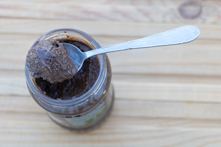 A spoonful of dark chocolate spread from an open glass jar on a light wooden table, close-up viewの写真素材