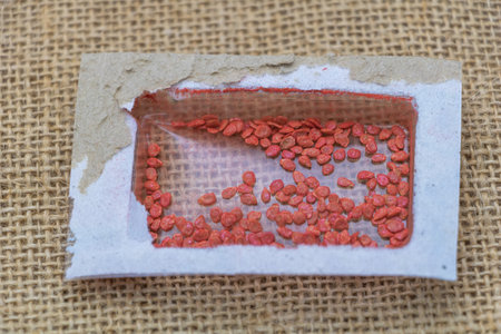 A small rectangular plastic seed packet with red seeds inside, on a burlap background.
