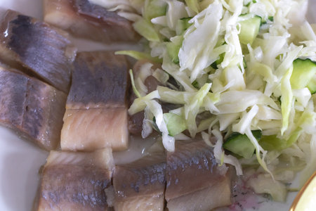 A close-up of sliced herring with cabbage and cucumber salad, arranged on the side of an open white plate. The fish is fresh from sea to table.の写真素材