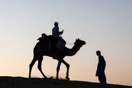 silhouetted against the bright orange hues of a dusk twilight, a single camel with rider is seen trotting away  past a male standing on the way side のeditorial素材