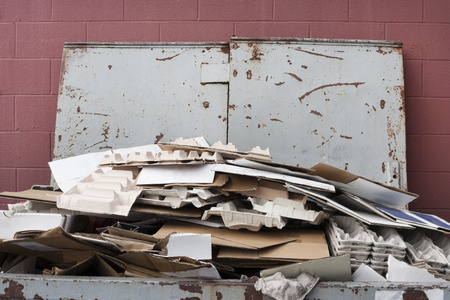 Close up of dumpster filled with cardboard for recycling. Lid open against reddish wall. Horizontal.