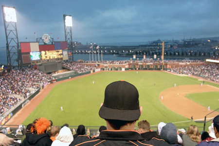 Baseball fan watching night game in San Francisco. The fan is wearing a black baseball cap turned backwards and a black jacket with orange piping. We can see the infield, outfield, stadium lights.の写真素材