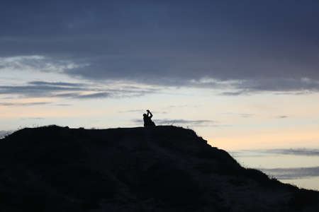 silhouette of a man in a cap sitting on a mountain watching the sunset.の写真素材