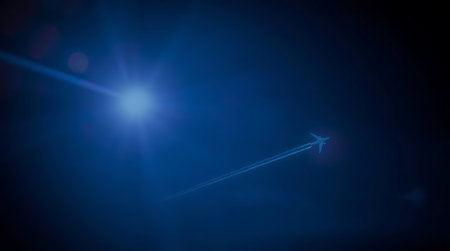 A distant airplane cuts through the deep blue sky, leaving a distinct vapor trail. A bright sun flare adds a dramatic, luminous effect to this scene of aerial journey and travel.の素材