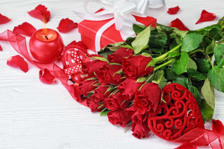 Valentine's Day background with a bouquet of beautiful red roses, gift box, heart shaped ornaments and globe shaped candle on white wooden table (selective focus).の写真素材