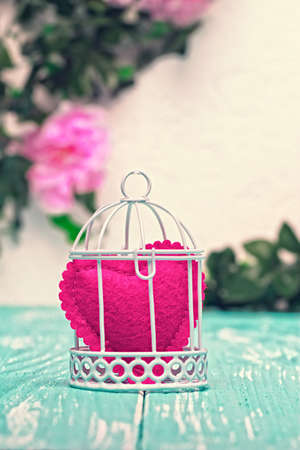Still life romantic background with heart shaped ornament in decorative bird cage on teal wooden planks and pink flowers on wall in the background (selective focus).の写真素材