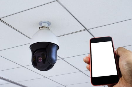 hand holding smartphone white display copy space with CCTV security camera installed wall ceiling, security camera system concept.の写真素材