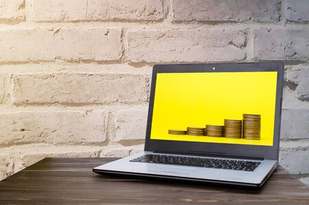 Laptop computer with  money coin stack growing on yellow background business in screen on wooden table with brick wall background, selective focus,  financial, business, saving money concept.の写真素材