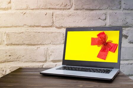 Laptop computer with red ribbon bow on yellow background in screen on wooden table with brick wall background, selective focus, copy space, sunlight effectの写真素材