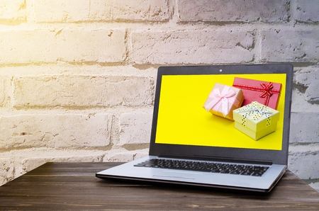 Laptop computer with colorful gift box on yellow background in screen on wooden table with brick wall background, holiday sale and online shopping concept, selective focus, copy spaceの写真素材