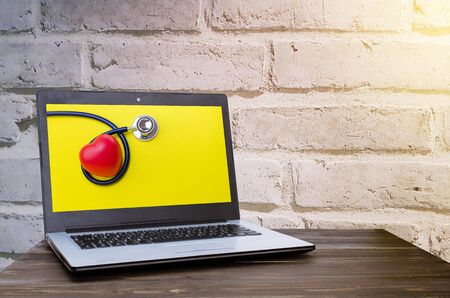 Laptop computer with red heart and stethoscope on yellow background in screen on wood table with brick wall background, selective focus, heart health care and medical technology conceptの写真素材