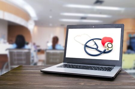 Laptop notebook computer with red heart and stethoscope on white background on wooden table with blurred hospital background, heart health care and medical technology concept, selective focusの写真素材