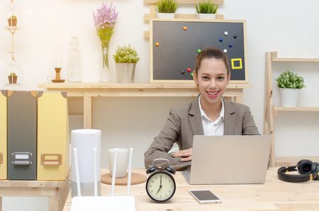 asian young business woman beautiful smile working on laptop notebook computer on wood table with alarm clock, headphone, smartphone, wireless router, document folder, selective focus,sunlight effectの写真素材
