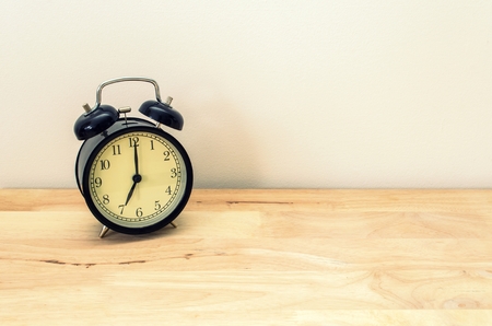 retro black color alarm clock times at 7 o'clock morning on wooden table table and white wall, vintage toneの写真素材