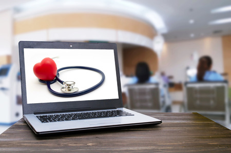 Laptop computer with red heart and stethoscope on white screen on wooden table with blurred view of patient sitting wheelchair in hospital background, heart health care and medical technology conceptの写真素材