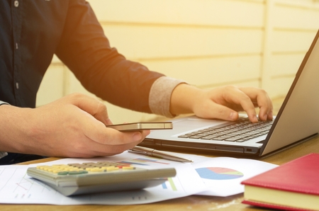 young accountant or business man using smartphone with calculator, data sheet, laptop on desk, analyzing and calculate, savings, finances, economy concept, sunlight effect, soft focus, selective focusの写真素材