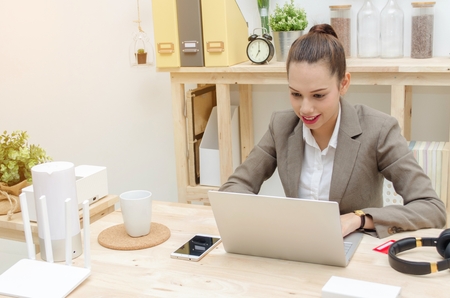 asian young business woman beautiful smile working on laptop notebook computer on wood table with alarm clock, headphone, smartphone, wireless router, document folder, soft focus,sunlight effectの写真素材