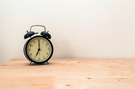 retro black color alarm clock times at 7 o'clock morning on wooden table table and white wall, vintage toneの写真素材