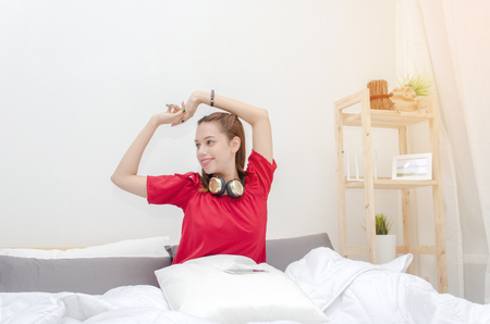 beautiful asian young woman in red pajamas with headphone on her neck and mobile smartphone waking up at the morning in bedroom, soft focus, good morning, holiday, lifestyle technology conceptの写真素材