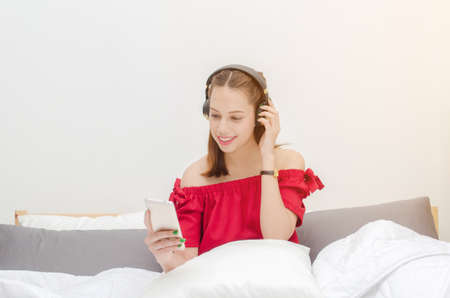 young asian smiling woman in red dress relaxing and using smartphone listening to music in headphone in bedroom at the morning, technology internet and holiday lifestyle concept, sunlight effectの写真素材
