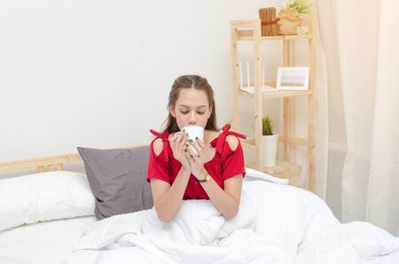 young asian woman beautiful smile in red pajamas with coffee cup on bed waking up at the morning in bedroom with sunlight effect, selective focus, soft focus, good morning, holiday, lifestyle conceptの写真素材