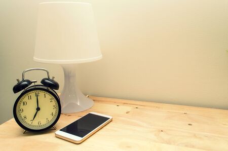 retro style black alarm clock, modern white lamp and mobile smart phone on wooden desk and white wall, vintage color tone, copy space, selective focusの写真素材